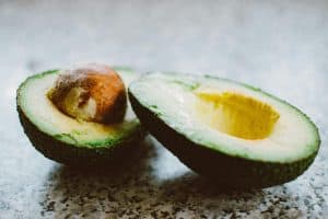 A ripe avocado, known as one of the top magnesium rich foods, is cut in half on a speckled surface. One half cradles the brown seed while the other reveals its creamy green flesh.