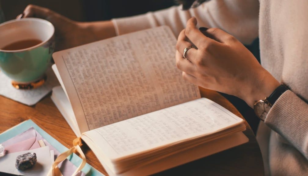 A person seeking better sleep holds a cup of tea and sits at a wooden table, reading or writing in an open journal with handwritten notes. Nearby are envelopes, a ribbon, and a few crystals.