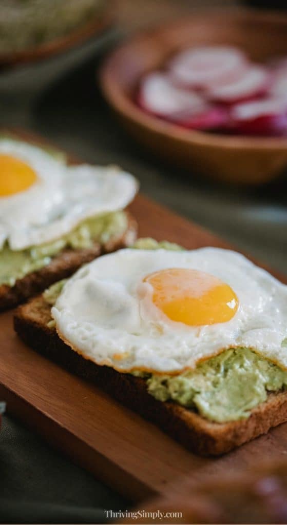 Two slices of toast topped with mashed avocado and sunny-side-up fried eggs are served on a wooden board—a perfect choice for busy people seeking nourishing morning routines. In the background, there’s a blurred dish with sliced radishes.