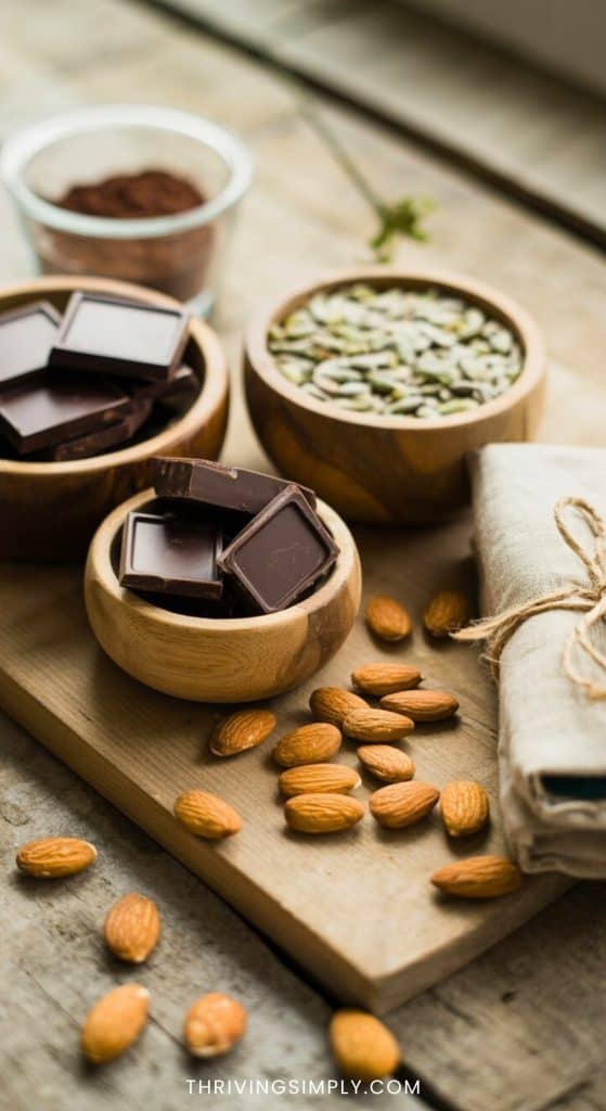 Wooden bowls filled with dark chocolate pieces and pumpkin seeds, both magnesium rich foods, sit on a wooden board beside whole almonds and a folded linen napkin tied with twine. A small glass dish of cocoa powder is in the background.