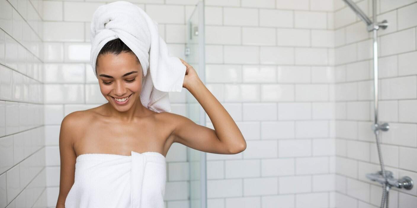A woman wrapped in a white towel stands in a white tiled bathroom, smiling with a towel around her hair, next to a glass shower door and wall-mounted showerhead, enjoying her calming ritual as part of her Gentle Everything Shower routine.