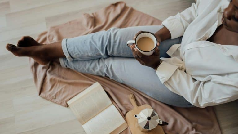 A person embracing slow living sits on a blanket with legs stretched out, holding a cup of coffee. An open book and moka pot rest nearby on the light wooden floor. The person wears light jeans and a white shirt.