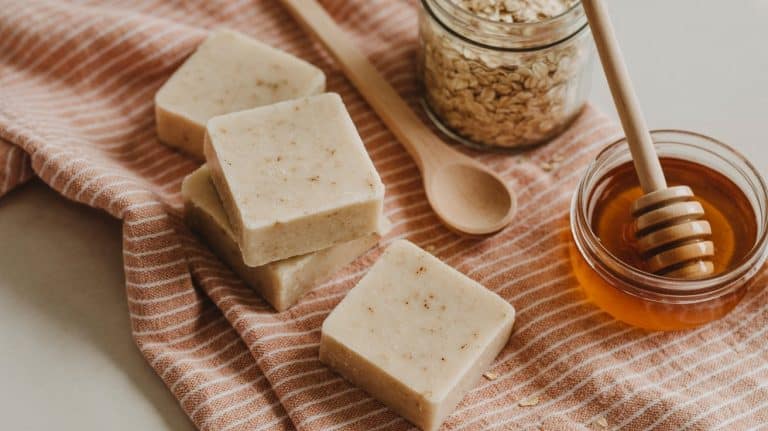 Three bars of handmade Oatmeal soap rest on a striped cloth beside a jar of oats, a wooden spoon, and a glass bowl of honey with a honey dipper, evoking a natural, homemade skincare vibe.