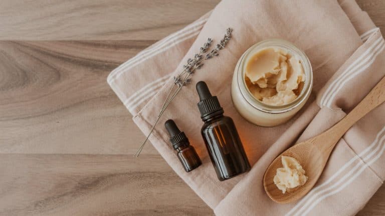 A jar of creamy balm, two brown dropper bottles, a wooden spoon with balm, and a sprig of lavender rest on a folded beige cloth atop a wooden surface.