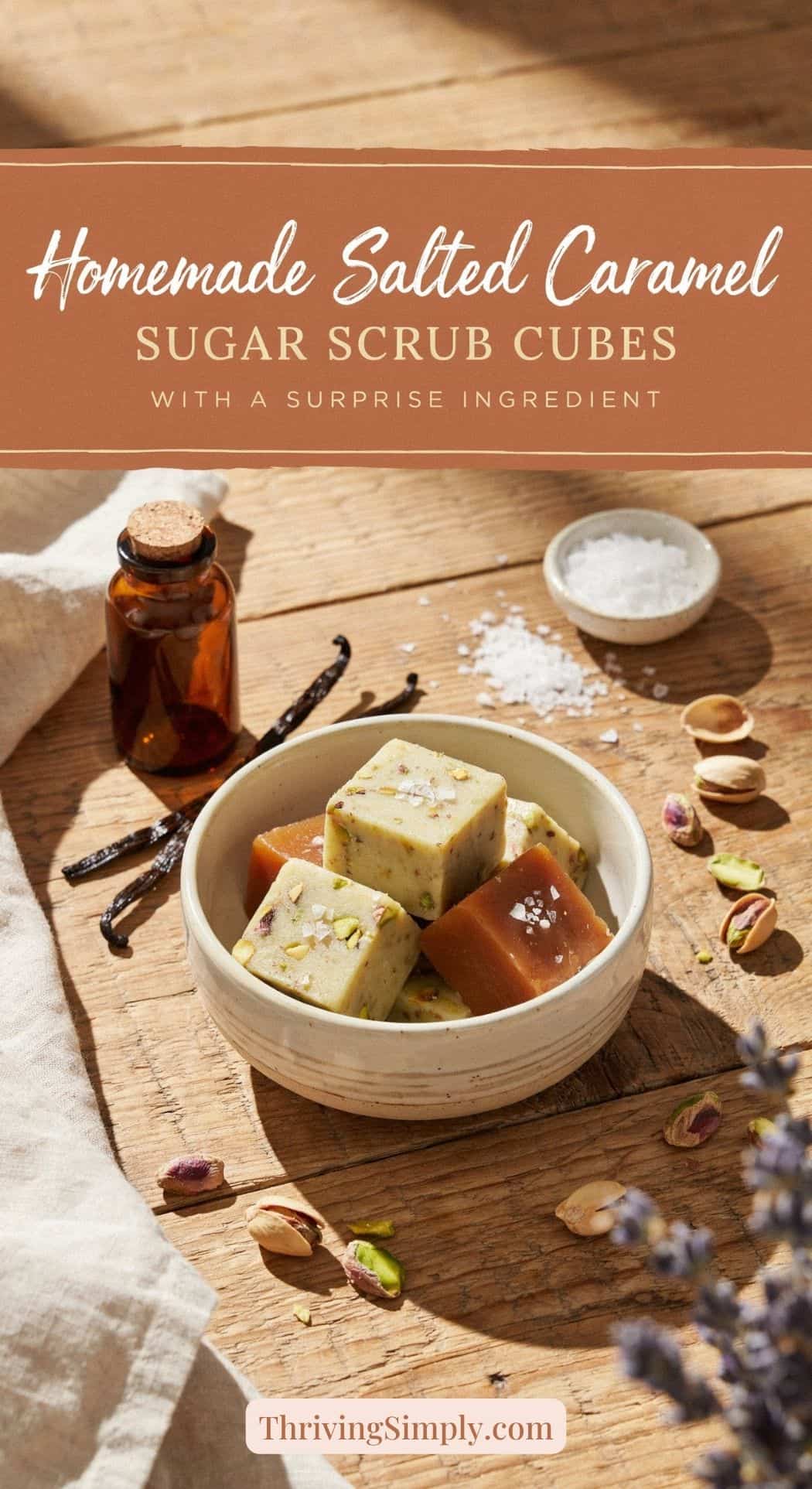 A bowl of homemade Salted Caramel Sugar Scrub Cubes sits on a table with pistachios, salt, a vanilla bean, an amber bottle, and purple flowers nearby. Text above reads, Homemade Salted Caramel Sugar Scrub Cubes.
